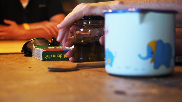 Ein Tisch mit verschiedenen Gegenständen, darunter ein Glas, eine Tasse und ein Kartenspiel. Im Hintergrund sind verschwommene Figuren zu sehen.