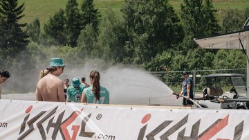Eine Gruppe von Menschen steht an einem Veranstaltungsort, während Wasser in die Luft gespritzt wird. Im Hintergrund sind grüne Bäume und eine Landschaft zu sehen.