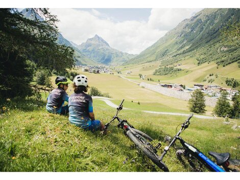 Zwei Kinder sitzen auf einer Wiese und blicken auf das Tal mit einem Dorf im Hintergrund. Neben ihnen stehen zwei Fahrräder, umgeben von einer schönen Berglandschaft.