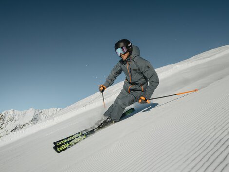 Ein Skifahrer fährt über eine schneebedeckte Piste in den Bergen. Im Hintergrund sind majestätische Berge und ein klarer Himmel zu sehen.
