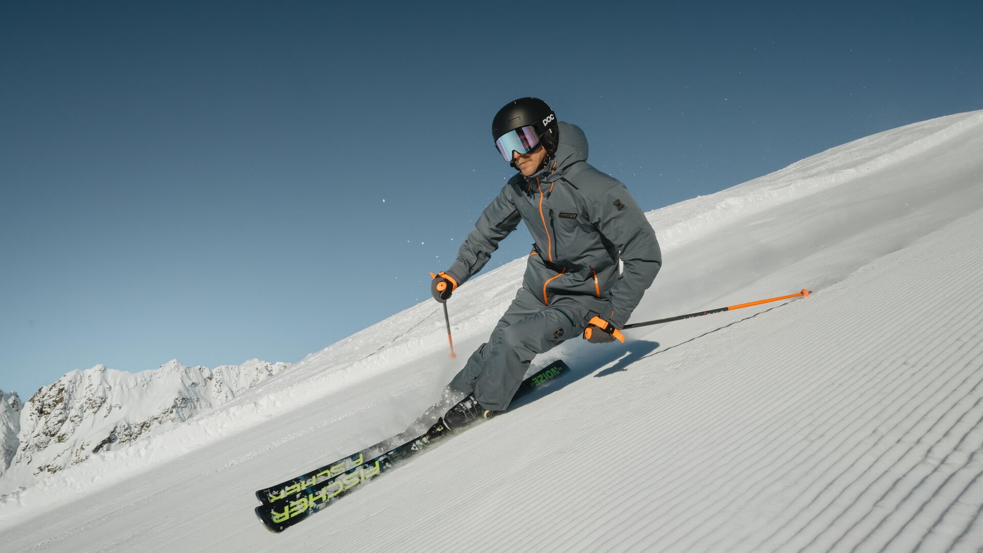 Ein Skifahrer fährt über eine schneebedeckte Piste in den Bergen. Im Hintergrund sind majestätische Berge und ein klarer Himmel zu sehen.