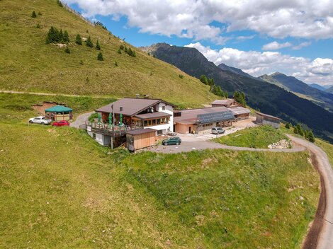 A mountain inn with several buildings and a large terrace in the midst of a green landscape. The surroundings are surrounded by mountains and a blue sky.