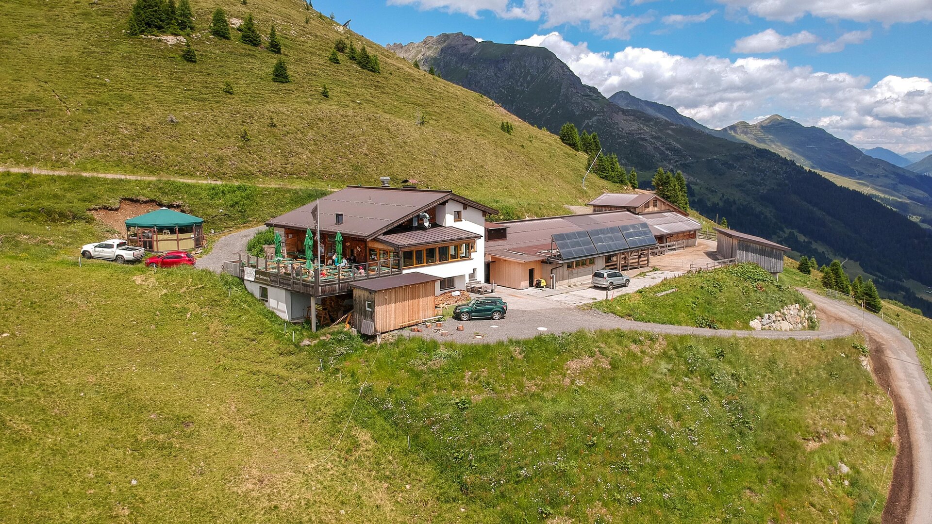 A mountain inn with several buildings and a large terrace in the midst of a green landscape. The surroundings are surrounded by mountains and a blue sky.