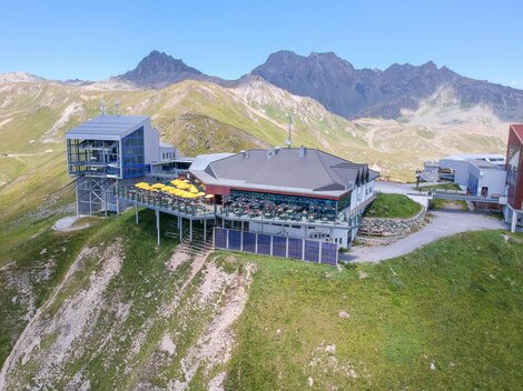 A modern mountain restaurant with a large terrace, surrounded by green meadows and mountains. The clear sky offers a dreamlike view of the alpine landscape.