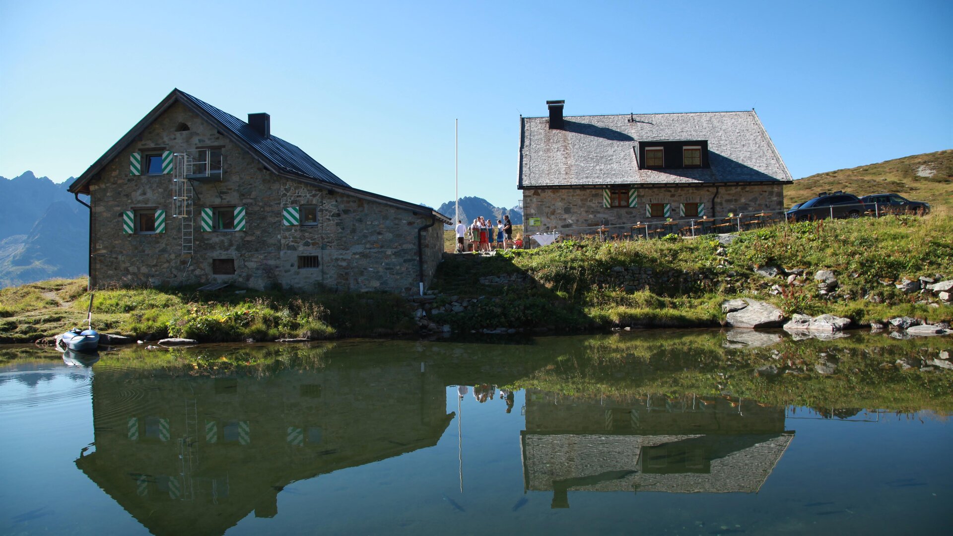 A picturesque mountain landscape with two cozy houses along a tranquil lake. The buildings reflect clearly in the water and are surrounded by green meadows.