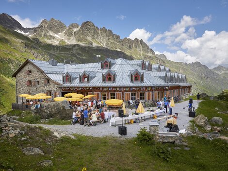 A charming mountain cabin with terraces and sun umbrellas. In the background, majestic mountains and a green meadow can be seen.