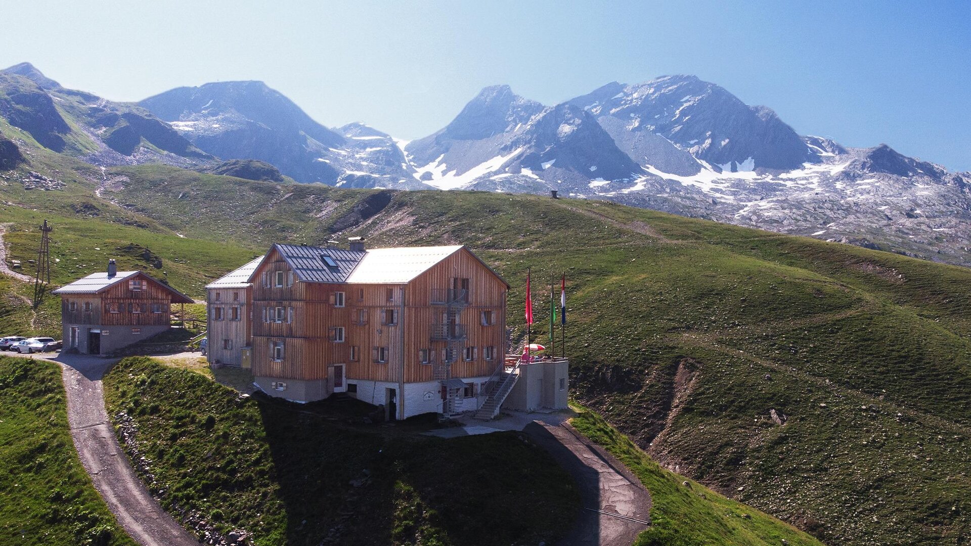 A modern wooden building is situated picturesque in the mountains. In the background, snow-covered peaks and a green landscape can be seen.