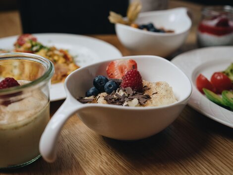 Ein schöner Tisch mit verschiedenen Frühstücksgerichten. Eine Schüssel mit Haferbrei, frischen Beeren und Schokolade ist im Vordergrund zu sehen.