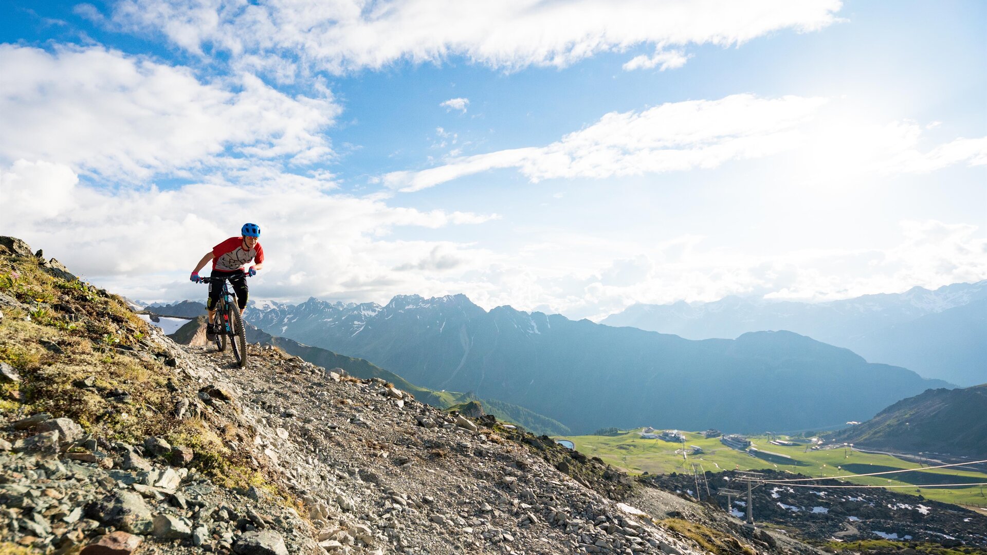 Ein Mountainbiker fährt über einen steilen, steinigen Pfad mit spektakulärer Berglandschaft im Hintergrund. Der Himmel ist klar und es gibt viele Wolken.