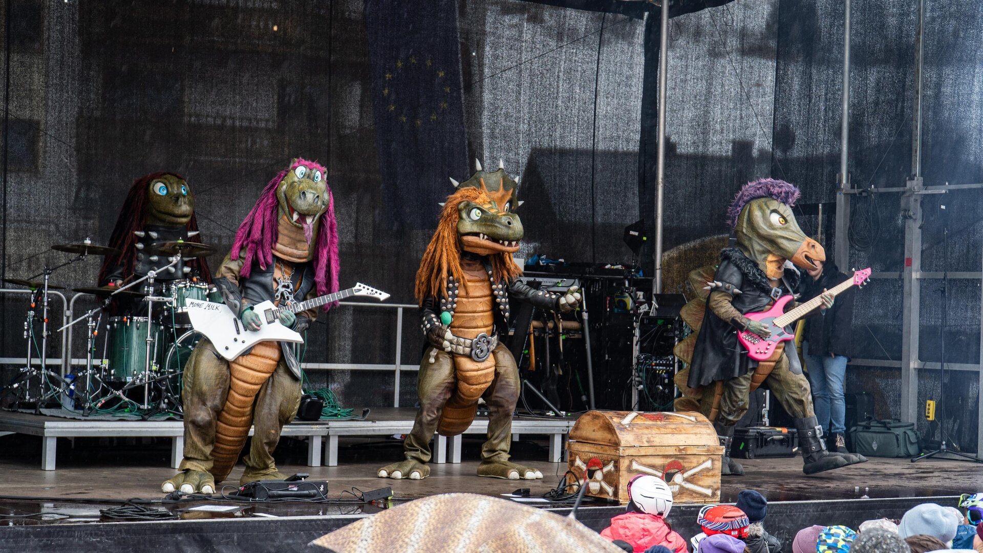 Eine Band von Drachen-Maskottchen spielt auf einer Bühne. Im Hintergrund sind Musikinstrumente und eine begeisterte Menge zu sehen.