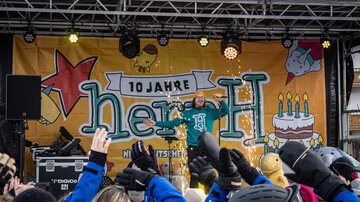 Ein Künstler steht auf der Bühne und begeistert das Publikum. Im Hintergrund sind Dekorationen für einen Jubiläumsfeier zu sehen.