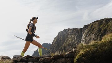 Eine Läuferin, die auf einem Bergpfad joggt, umgeben von beeindruckenden Felsen und einer weiten Aussicht. Sie trägt sportliche Kleidung und hat einen Gehstock in der Hand.