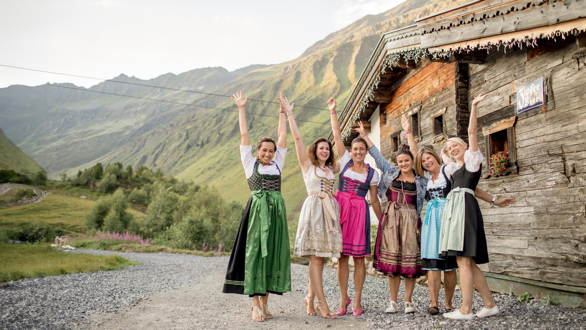 Sechs Frauen in traditioneller Tracht stehen fröhlich vor einem alten Holzhaus in den Bergen. Die Landschaft ist grün und hügelig mit einer beeindruckenden Bergkulisse.