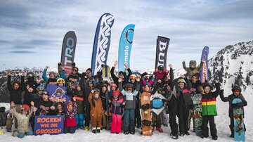 Eine Gruppe von Kindern und Erwachsenen steht fröhlich im Schnee. Sie tragen Skiausrüstung und feiern mit Fahnen im Hintergrund.