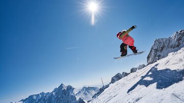 Ein Snowboarder springt über eine schneebedeckte Piste. Der Himmel ist blau und die Sonne strahlt hell.