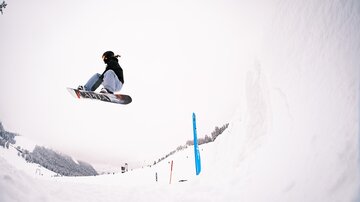 Ein Snowboardfahrer springt hoch in der Luft über einen verschneiten Hang. Im Hintergrund sind Bäume und eine winterliche Landschaft zu sehen.