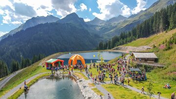 Eine lebhafte Veranstaltung in den Bergen mit vielen Menschen, die sich um einen klaren See versammeln. Bunte Zelte und eine schöne Naturlandschaft umgeben die Szene.