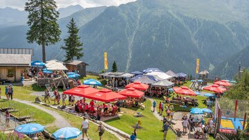 Eine lebhafte Berghütte mit Terrassen unter roten und blauen Sonnenschirmen. Im Hintergrund sind beeindruckende grüne Berge und ein klarer Himmel zu sehen.