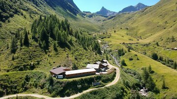 Eine malerische Berglandschaft mit einem ruhigen Tal und grünen Wiesen. Ein kleiner Bauernhof liegt entlang einer kurvigen Straße, umgeben von majestätischen Bergen.