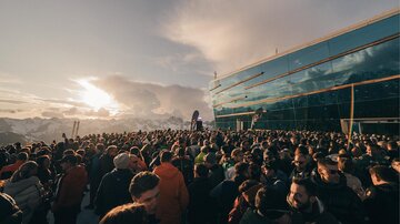 Eine große Menschenmenge versammelt sich vor einem futuristischen Gebäude in den Bergen. Im Hintergrund sind schneebedeckte Gipfel und ein Sonnenuntergang zu sehen.