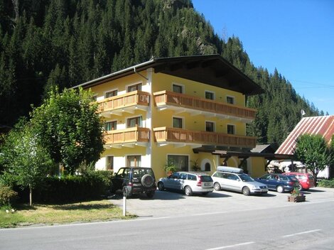 Ein helles Gebäude mit mehreren Balkonen, umgeben von Bäumen und Bergen. Auf dem Parkplatz stehen mehrere Autos.