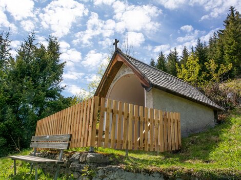 Eine kleine Kapelle mit einem Holzzaun steht in einer bewaldeten Umgebung. Der Himmel ist teilweise bewölkt und die Landschaft wirkt ruhig und friedlich. | © Bernhard Gruber – Photography