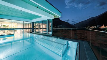 A modern swimming pool with smooth water on a terrace. In the background, the mountains and the evening sky are visible.