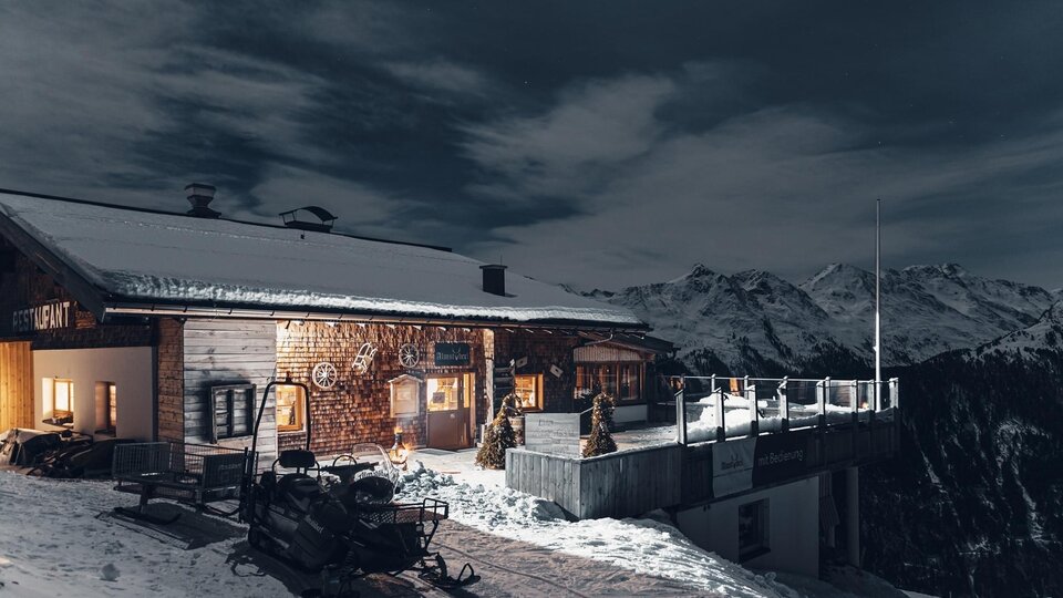 Eine Holzski-Hütte im Schnee mit einem atemberaubenden Bergpanorama. Die Szenerie ist bei Nacht mit einem klaren Himmel und einem beleuchteten Innenbereich.