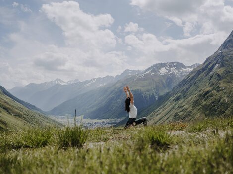 Eine Person übt Yoga in einer malerischen Berglandschaft. Die Umgebung ist grün und die Berge sind teilweise schneebedeckt, unter einem klaren Himmel.