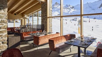 Ein modernes Restaurant mit großen Fenstern, das einen Blick auf eine verschneite Ski-Piste bietet. Die Einrichtung ist stilvoll und einladend, ideal für eine Pause nach dem Skifahren.