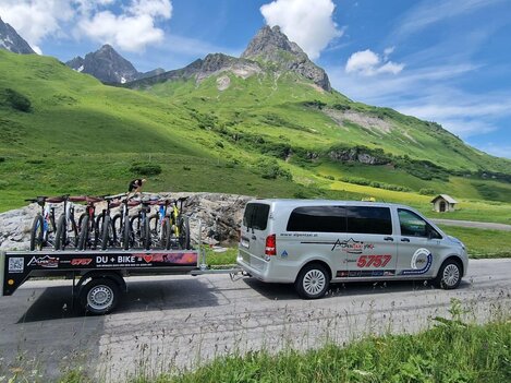 A vehicle with a trailer full of bicycles drives through a green mountain landscape. The surroundings are sunny with impressive mountains in the background.