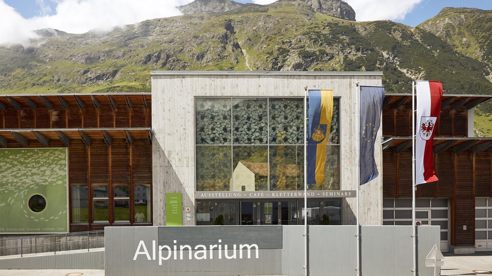 Ein modernes Gebäude des Alpinariums mit großen Fenstern. Im Hintergrund sind grüne Berge und ein blauer Himmel zu sehen.