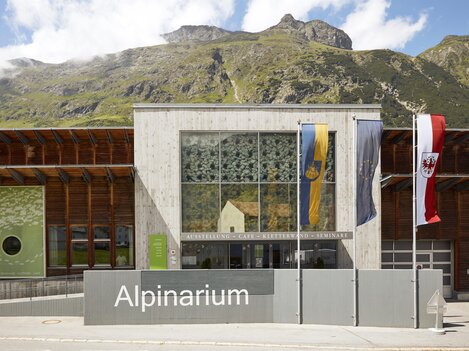 A modern building of the Alpinarium with large windows. In the background, green mountains and a blue sky can be seen.