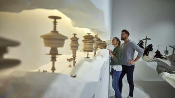 Ein Paar steht vor einer Kunstinstallation mit skulpturalen Objekten auf einer hellen Wand. Die Atmosphäre ist modern und inspirierend. | © TVB Paznaun - Ischgl