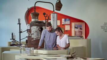 Ein Paar betrachtet Ausstellungsstücke in einem Museum. Im Hintergrund sind historische Objekte und informative Tafeln zu sehen. | © TVB Paznaun - Ischgl