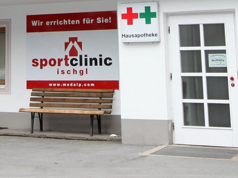 Eine Sportklinik mit einem roten Schild und einer Bank im Freien. Nebenan befindet sich ein Apothekenzeichen.