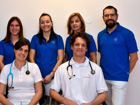 Ein Team von Fachleuten steht zusammen in blauen T-Shirts. Zwei Personen tragen Stethoskope und lächeln freundlich in die Kamera.