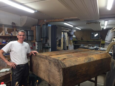 Ein Handwerker steht in seiner Werkstatt neben einer großen, alten Holzkiste. Im Hintergrund sind verschiedene Werkzeuge und Maschinen zu sehen.