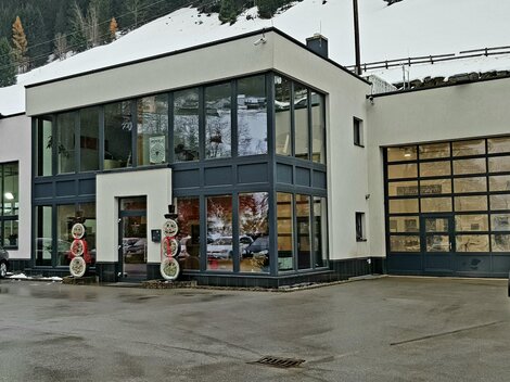 Ein modernes Autohaus mit großen Fenstern und einer modernen Architektur. Vor dem Gebäude stehen mehrere Fahrzeuge und im Hintergrund sind schneebedeckte Berge zu sehen.
