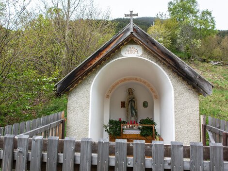Eine kleine Kapelle mit einem heiligen Standbild steht in der Natur. Sie ist von einem Holzzaun umgeben und von Bäumen umgeben. | © Bernhard Gruber – Photography