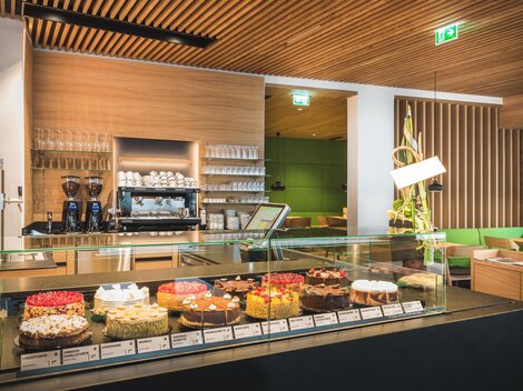 Eine moderne Café-Theke mit einer Auswahl an verschiedenen Kuchen. Im Hintergrund sind Holzverkleidungen und eine gemütliche Sitzgelegenheit sichtbar.