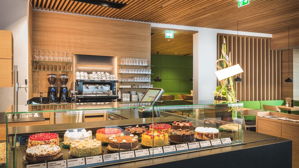 A modern café counter with a selection of various cakes. In the background, wooden paneling and a cozy seating area are visible.