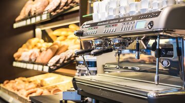 A modern espresso machine stands in front of a shelf with fresh rolls. In the background, various baked goods are clearly visible.