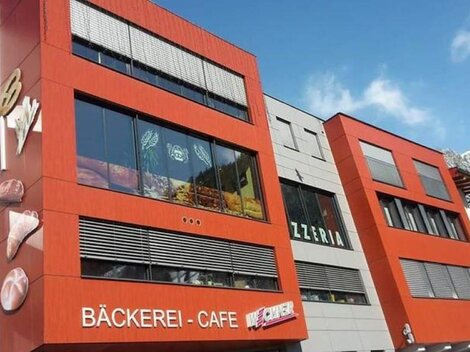 Ein modernes Gebäude mit auffälliger roter Fassade. Im Erdgeschoss befinden sich eine Bäckerei und ein Café.