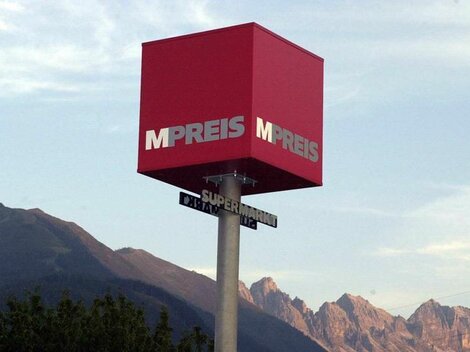 Ein rotes Werbeschild mit der Aufschrift "MPREIS" steht vor einem Gebirgshorizont. Der Schild zeigt auf eine Straße mit dem Zusatz "Supermarkt".