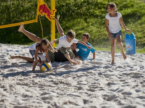 Eine Gruppe von Kindern spielt begeistert Volleyball im Sand. Zwei Kinder liegen im Sand, während die anderen um den Ball herum spielen.