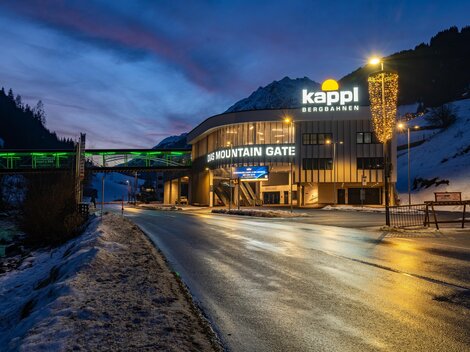 Ein modernes Gebäude mit dem Schriftzug "Kappl Bergbahnen" steht im Vordergrund. Die Szenerie zeigt eine winterliche Umgebung bei Dämmerlicht.