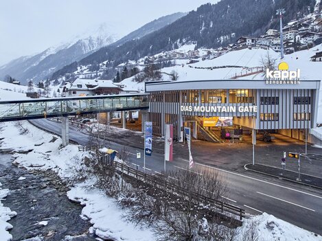 A modern building with the inscription "THE MOUNTAIN GATE" stands in a snow-covered landscape. In the background, mountains and a river are visible.