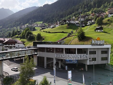 Ein modernes Gebäude mit der Aufschrift "Kappl" in einer bergigen Landschaft. Im Hintergrund sind grüne Hügel und kleine Häuser zu sehen.