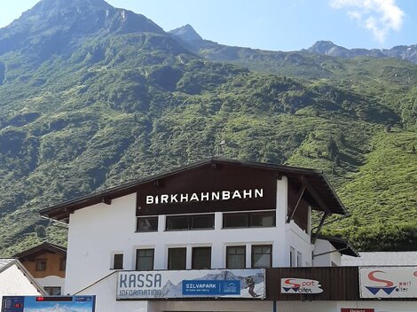Eine Bergbahnstation mit dem Namen "Birkhahnbahn" befindet sich im Vordergrund. Im Hintergrund sind grüne Berge und ein blauer Himmel zu sehen.